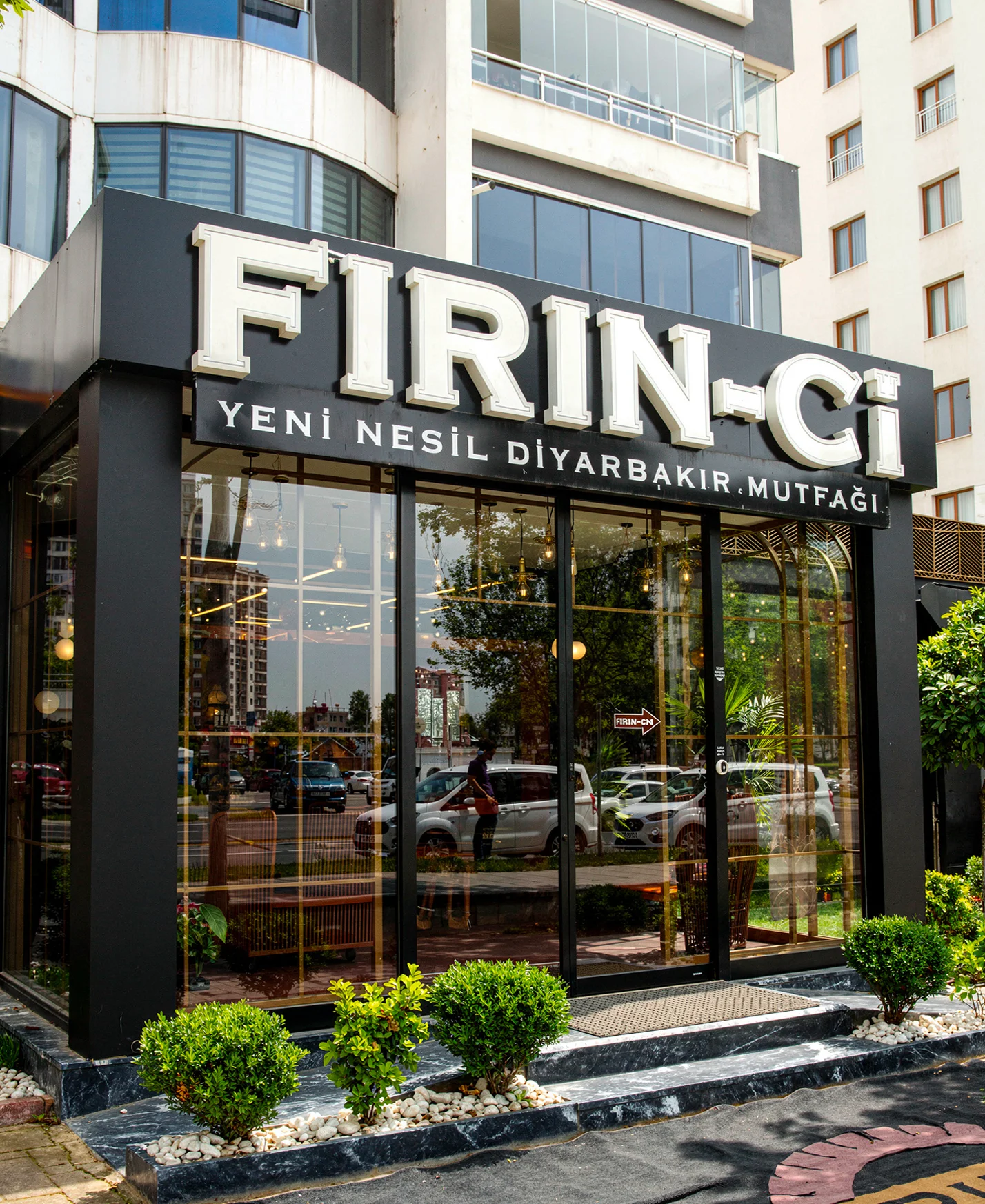 Koyu renk ahşap kaplama kavisli restoran bankosu, pirinç detaylı dairesel duvar apliği ve geniş salon bitkisi, NDO Mimarlık iç mimari projesi