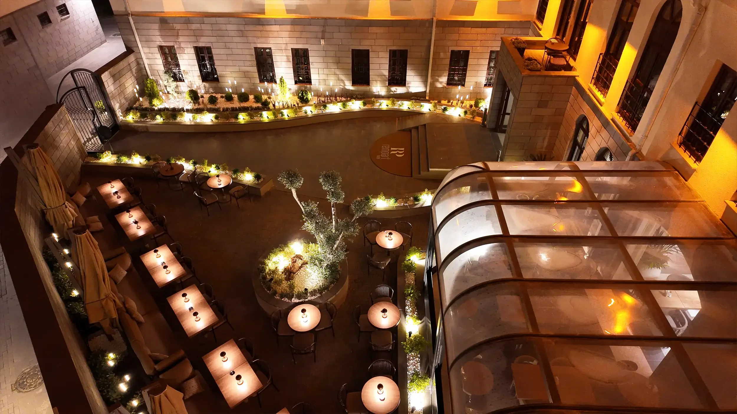 Rempart Hotel gece atmosferinde duvar diplerinden yükselen sıcak ışıklar, bitki adaları, masa düzenleri ve tarihi avlu duvarlarıyla oluşturulan butik otel dış mekânı, NDO Mimarlık